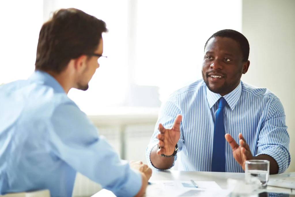 Two businessmen in discussion at a table, one gesturing with hands.