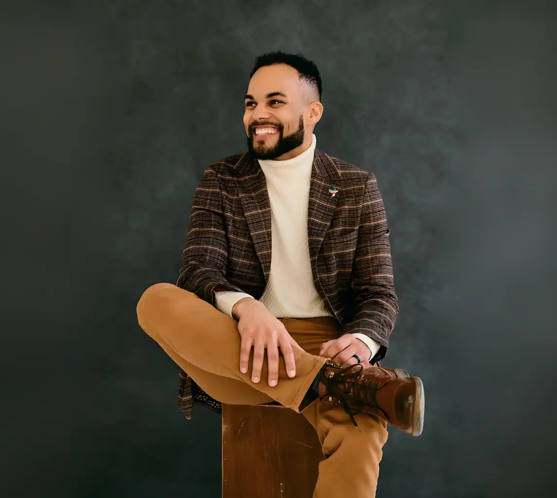 Man in stylish outfit smiling, seated with crossed legs against dark backdrop.
