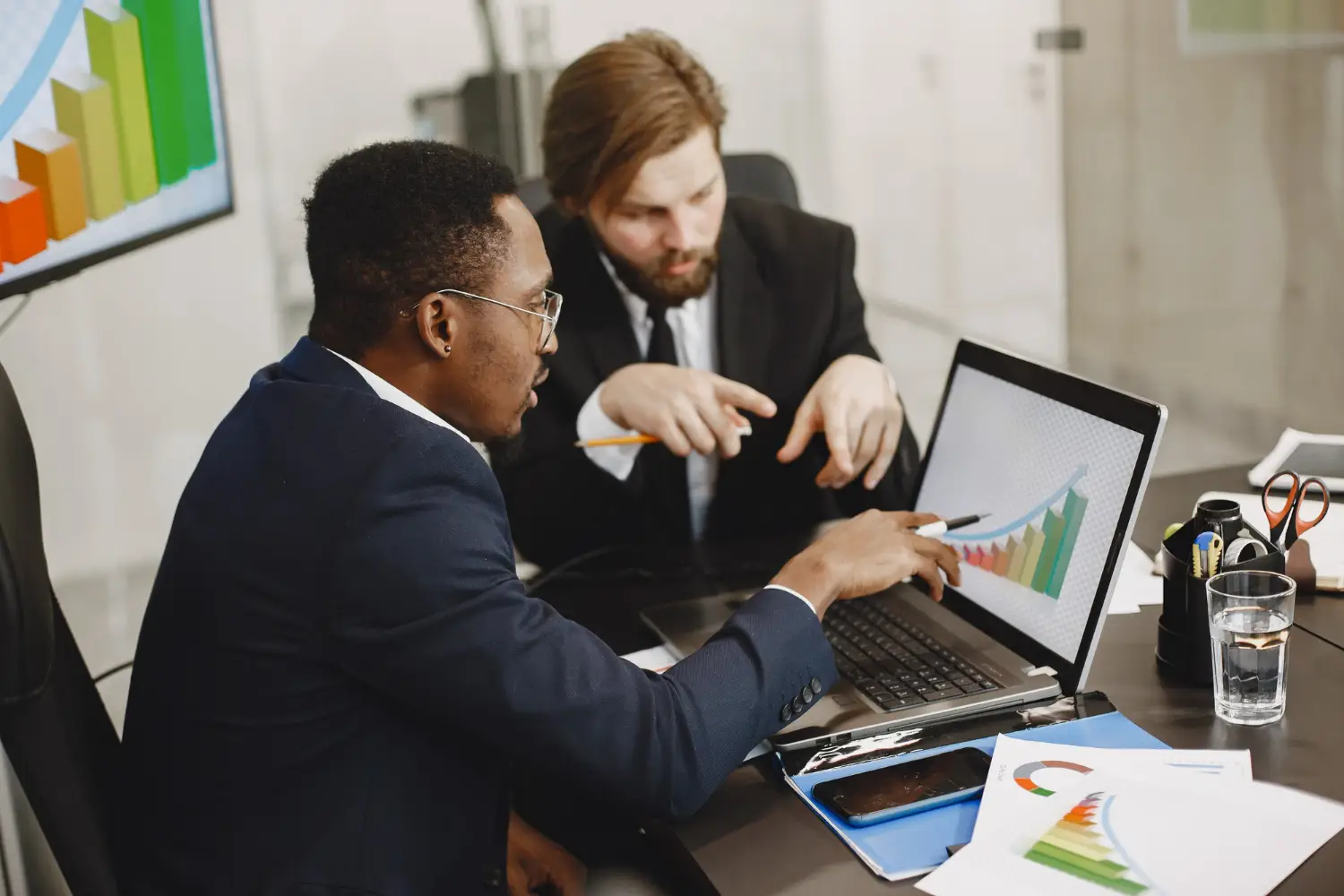 Two businessmen analyzing graphs together on a laptop during a meeting.