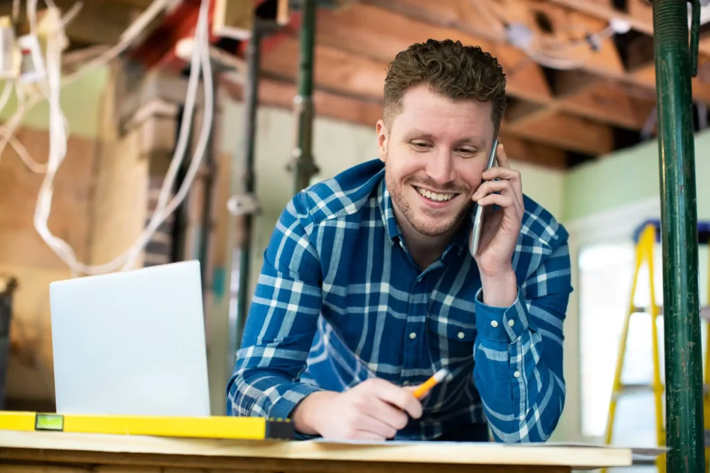 Man smiling while talking on the phone, working at a desk with a laptop and a pencil in a construction setting, representing business planning and advisory services in Saskatchewan.