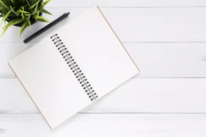 Open blank notebook with pen and plant on white wooden desk.